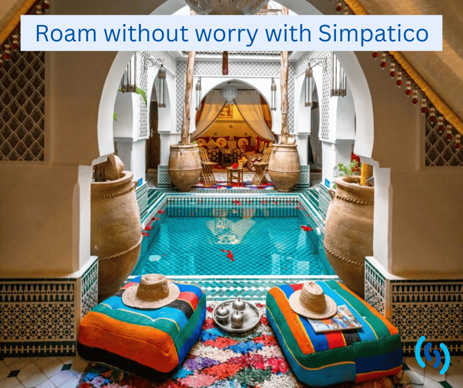 An image of a Moroccan Spa with a clear blue pool in the centre which has red rose petals floating on the surface. In the foreground are 2 brightly coloured cushions with straw hats positioned on the top of each. In the background are more cushions, chairs and drapes