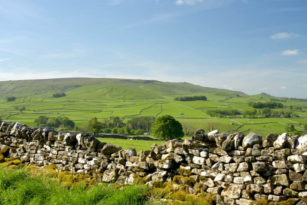 Hi there, this is a very scenic image composed of a hazy blue sky above some rolling green hills on which you can see all the farmer’s dry stone walls segmenting the fields. There’s a couple of remote houses dotted about, and in the foreground is a close up of a dry stone wall.