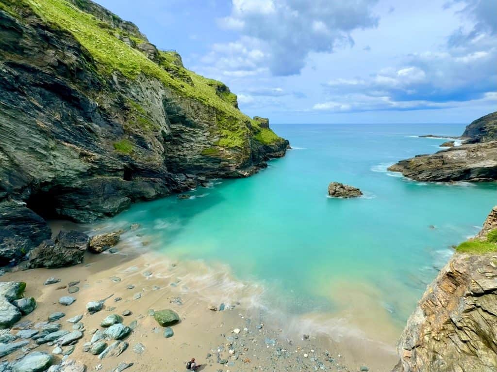 Hi there, this is an image taken in Cornwall using an iPhone 15 Pro Max. The image is a scenic shot of a little beach cove in Cornwall, in the foreground is a rocky/sandy beach surrounded on both sides by rugged cliffs. The sea is a clear turquoise blue, and above that is a typical blue sky with a few clouds dotted around.