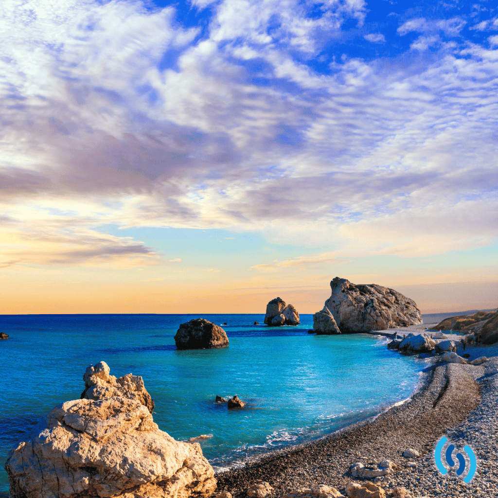Hello this is an image of a beach in Cyprus. The sea is clear and blue, the beach appears to be shingle with some rocky outcrops. The sky is a hazy blue and orange as if the sun is about to set, with some wispy clouds that look like they are drifting off into the distance. In the bottom left corner is the Simpatico logo.