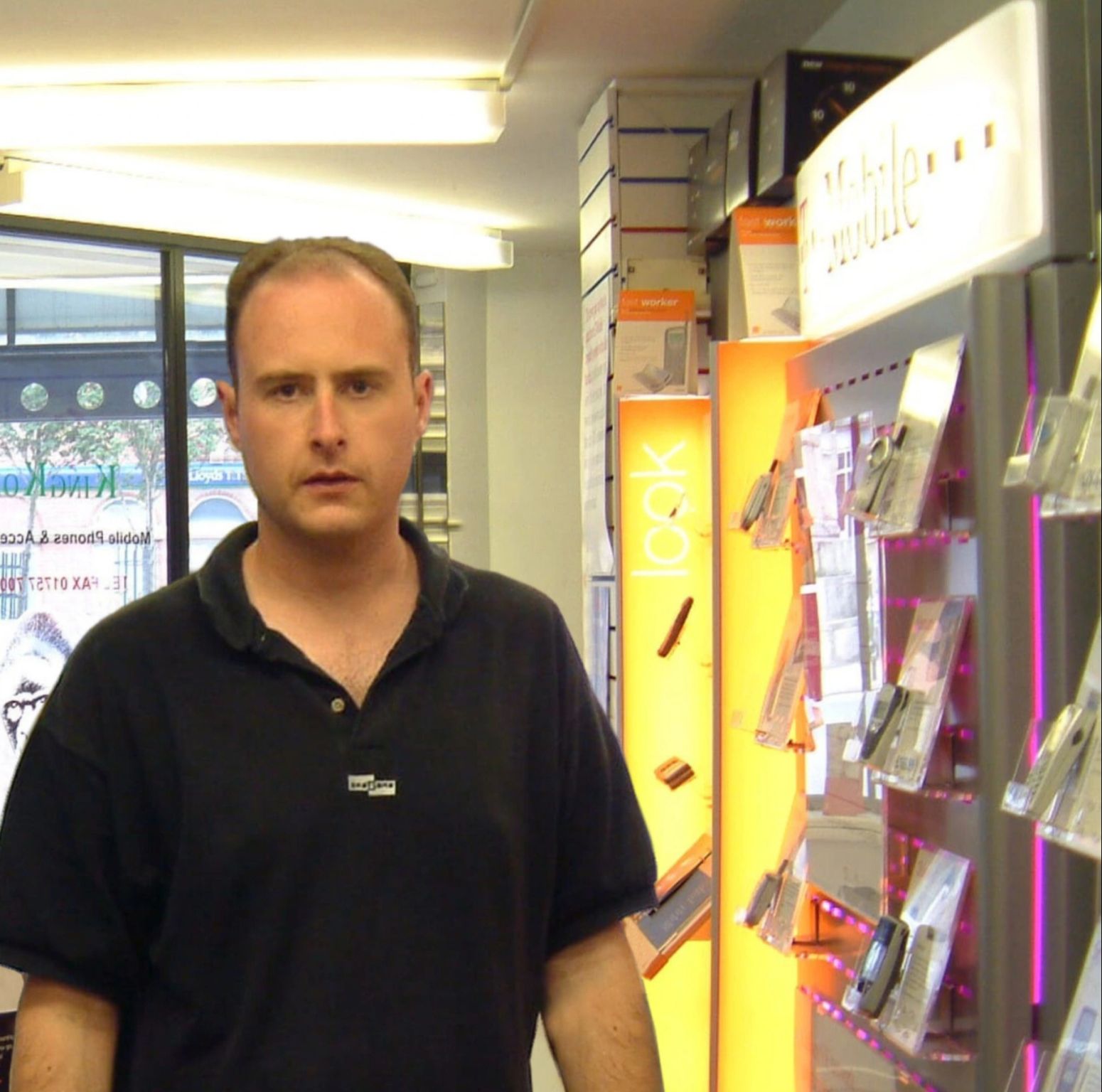 Dave is a young male wearing a black t-shirt and with a receding hairline! He is stood in a mobile phone shop from the late 1990s