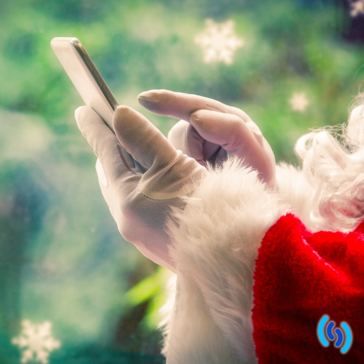 Christmas image with green foliage and snowflakes in the background. In the foreground, there is a Father Christmas's hand holding a mobile phone