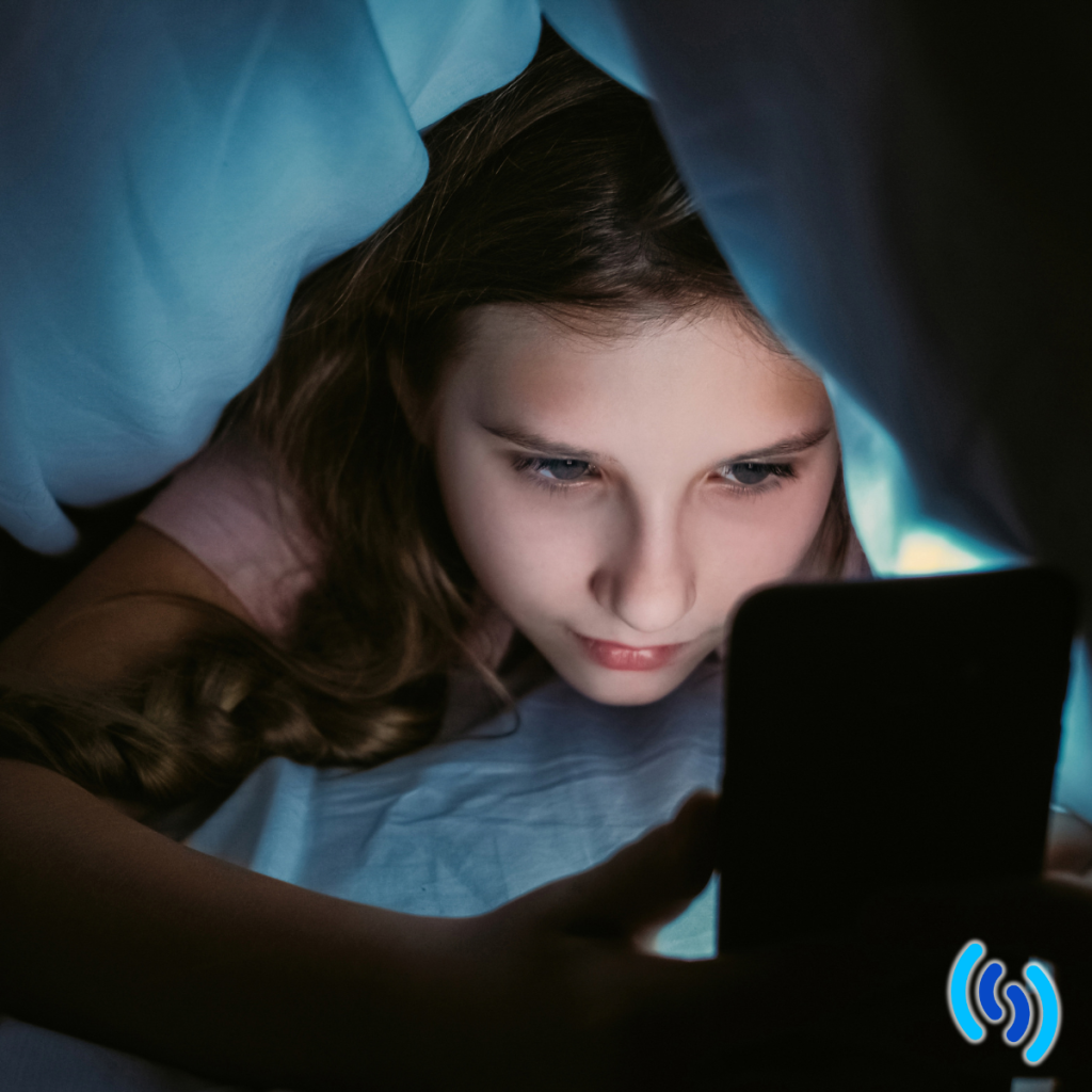 Very dark image of a teenage girl under the covers looking at the glowing screen of her smartphone